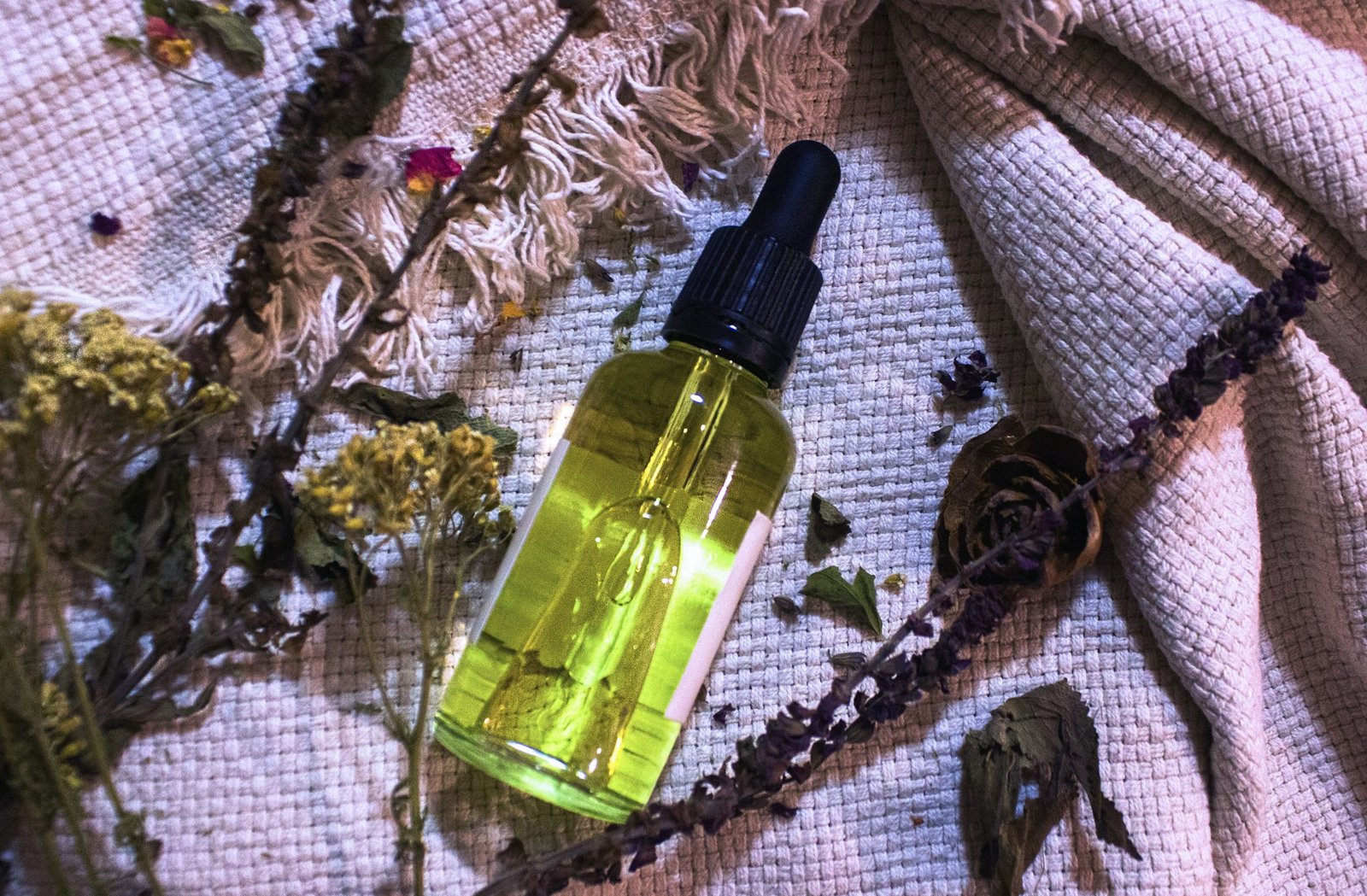 A dropper bottle of golden botanical oil surrounded by dried herbs and flowers