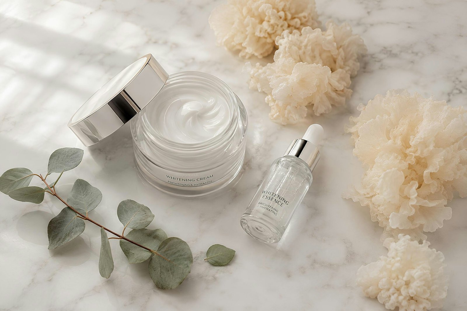 An open jar of white moisturizer cream next to a serum bottle and eucalyptus on marble